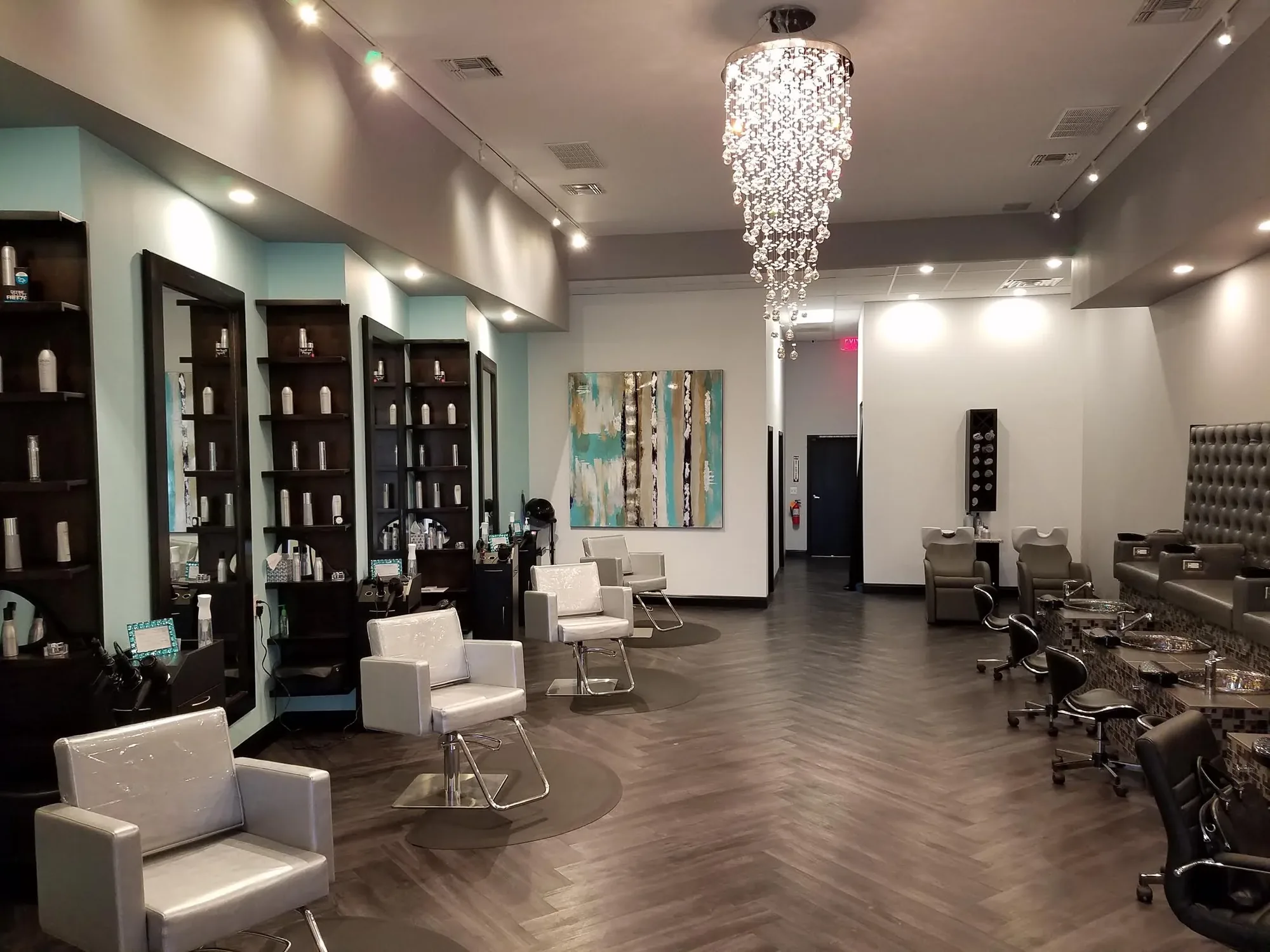 The Beauty Lounge salon floor: white styling chairs, dark mirror stations and a large crystal chandelier over a herringbone floor.