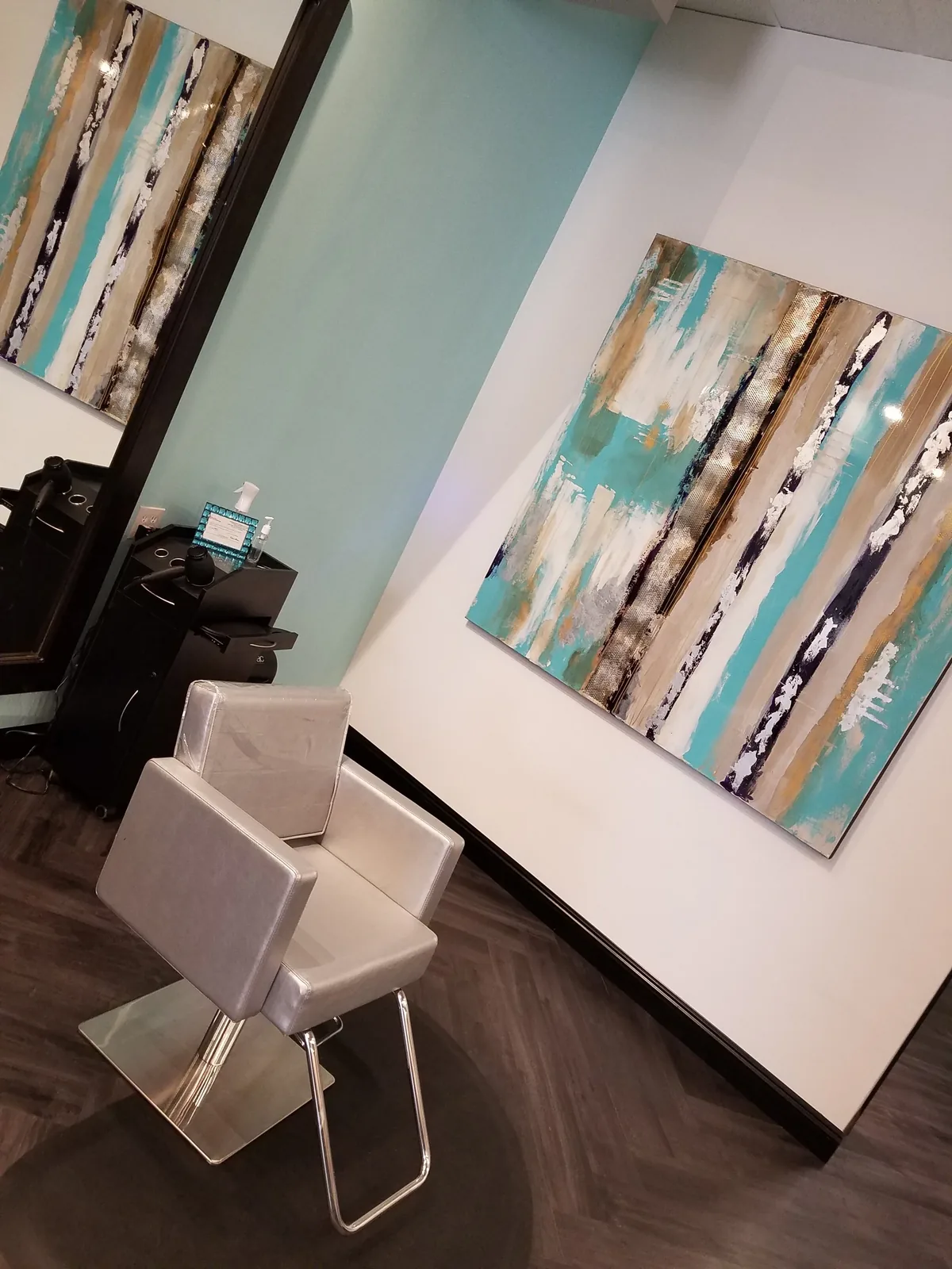 A silver styling chair beside a teal, gold and white abstract painting on an aqua accent wall.
